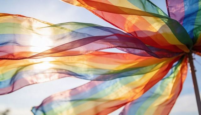 Backlit rainbow streamers fluttering under blue sky, sunlit translucent pride fabric motion