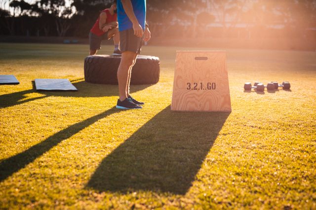 Low section of a fit Caucasian man standing next to a wooden box with '3, 2, 1, GO' written on it, during an outdoor cross training session at sunset. Various training equipment, including a large tire and dumbbells, are scattered on the grass. This image is ideal for promoting fitness programs, outdoor exercise routines, and healthy lifestyles.