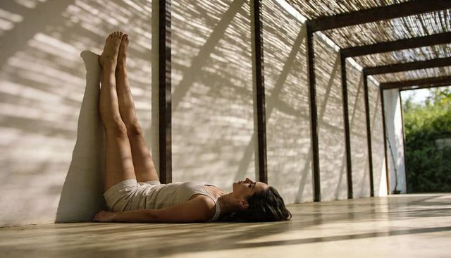 Woman resting with legs up against white wall under pergola casting linear sun shadows