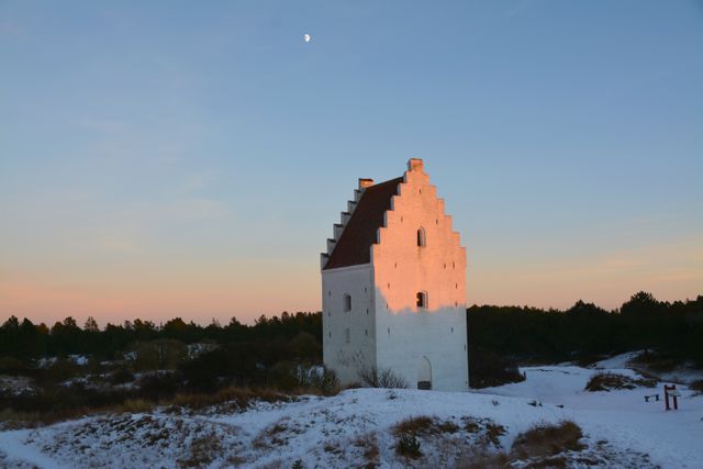 Abandoned white church standing solemnly in a snowy landscape, illuminated by the warm light of sunset. The sky is clear with a visible crescent moon, contributing to the serene ambiance. Suitable for use in topics related to historical architecture, rural scenes, winter scenery, or exploring themes of solitude and tranquility.