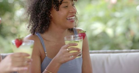 Laughing friends sipping strawberry-garnished citrus cocktails on sunlit patio sofa