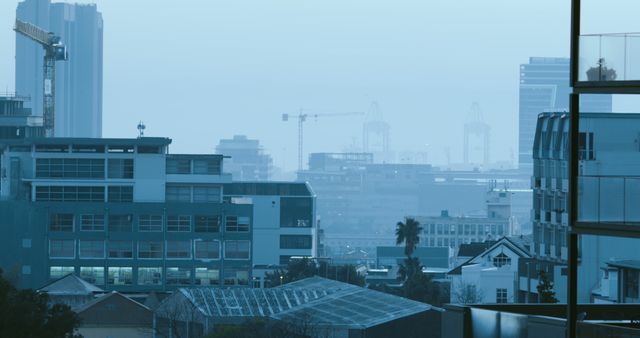 Mist covers early morning urban cityscape, construction cranes in distance. Ideal for conveying development, urban growth, city atmosphere, or architectural topics. Useful for articles, blogs, urban studies, marketing materials.
