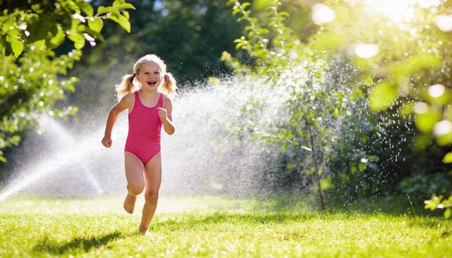 Joyful little girl running through backyard sprinkler in pink swimsuit on sunny summer day