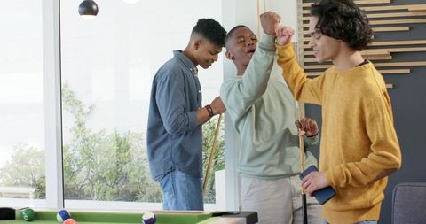 Diverse young men celebrating and playing pool in modern casual lounge with large windows