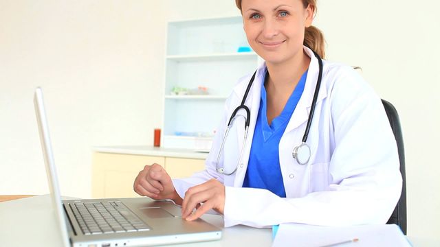 Female doctor is smiling while typing on a laptop, wearing a white coat and stethoscope, in a modern clinic setting. Ideal for use in healthcare advertisements, medical articles, blogs on healthcare technology, and hospital websites.