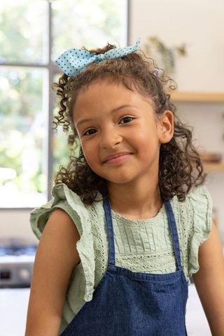 Smiling girl wearing denim apron and polka-dot bow in cozy sunlit home kitchen portrait
