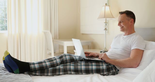 Man in pajamas working remotely from a bed using laptop. Suitable for depicting work-from-home culture, telecommuting, home offices, modern convenience, and the blending of home and work life.