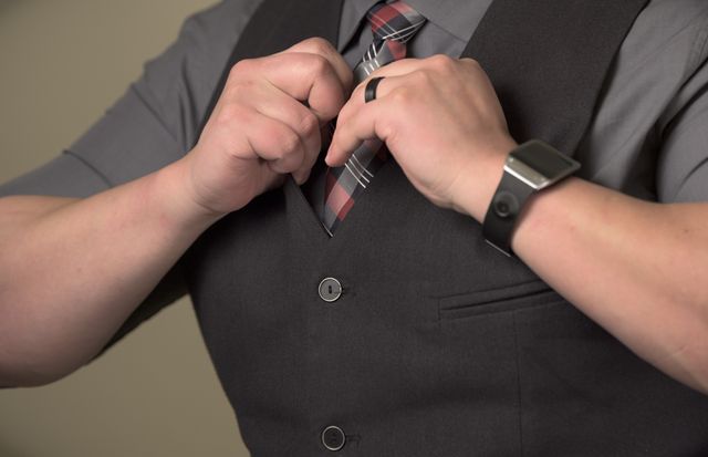This photo shows a man adjusting his tie while wearing a grey vest and a smartwatch. It is suitable for illustrating themes of business preparation, professional fashion, modern workplace trends, and corporate events. Useful for articles, blog posts, or advertisements focused on men's formal clothing, tech in fashion, or business attire standards.