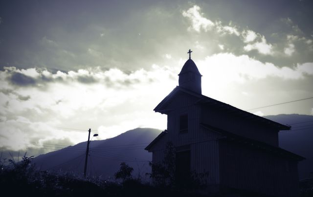 Silhouette of old church building against dramatic cloudy sky with soft evening light. Ideal for spiritual and religious themes, dramatic architectural references, or use in landscape and scenic contexts.