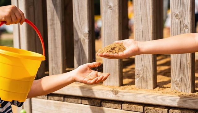 Children sharing sand while pouring from yellow bucket in sunny playground