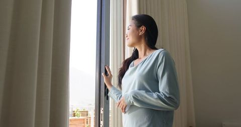Asian woman standing by sliding door looking out at balcony in serene natural daylight