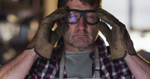 Image shows a tired construction worker wearing safety goggles and adjusting them in a dimly lit workshop. He is wearing heavy work gloves and a plaid shirt. The image captures a candid moment that portrays fatigue and dedication often seen in physical labor jobs. This can be used in articles or advertisements related to labor workforce, occupational safety, workwear, and construction industry awareness campaigns.