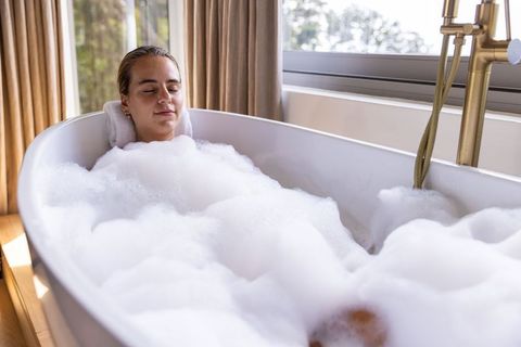 Relaxing woman soaking in freestanding bathtub with thick bubbles and warm sunlight