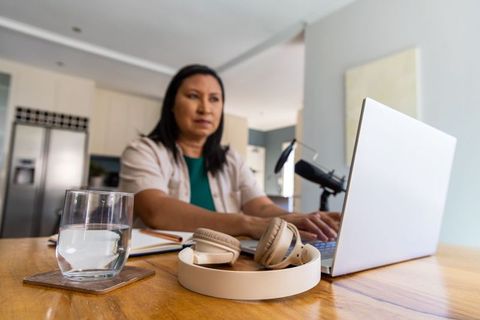 Asian woman working from home typing on laptop with podcast microphone and headphones