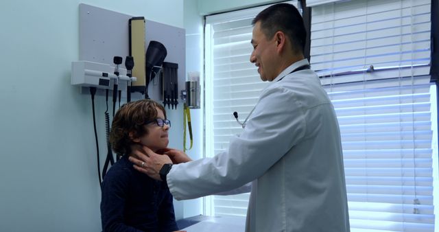 Pediatric doctor examining young boy in a clinic. Boy is sitting on a table while doctor checks his throat. Medical tools and diagnostic equipment visible on the wall behind. Ideal for content related to children's healthcare, medical clinics, pediatric care, health checkups, and doctor-patient interactions.