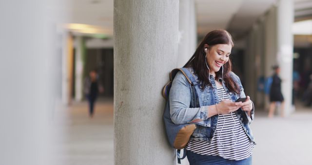 This image shows a young woman leaning against a column, smiling while using a smartphone. She appears relaxed, wearing casual clothing including a denim jacket and striped shirt. Earphones suggest she is listening to music. This image can be used in contexts such as lifestyle blogs, technology advertisements, urban living promotions, youth culture content, or digital age-related articles.