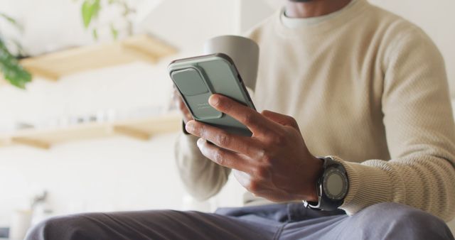 A man holding a smartphone and a mug in a modern home interior. He is dressed casually and appears to be sitting while using his phone. This image could be used to represent everyday life, technology usage, remote work, or casual leisure moments at home. Ideal for articles about mobile devices, technology integration in daily life, or advertisements for home lifestyle products.