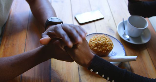 Two African American individuals are holding hands over a wooden table, with copy space. A sense of companionship or support is conveyed as they share a moment together with a coffee and muffin nearby.