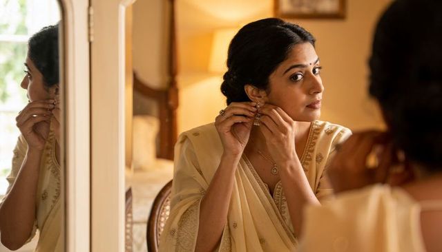Woman adjusting earrings in cream gold-trimmed saree at mirror during bridal preparation