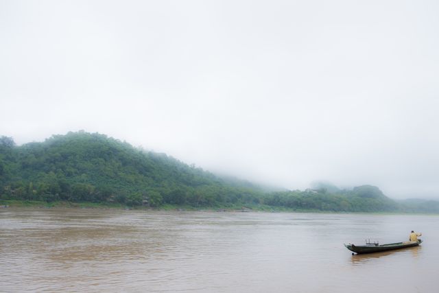 A lone fisherman is on a boat navigating a wide, peaceful river cloaked in dense fog. The mist envelops the lush green hills in the background, contributing to a serene and tranquil atmosphere. This image can be effectively used in travel brochures, environmental conservation campaigns, and advertisements promoting peaceful and picturesque rural destinations.