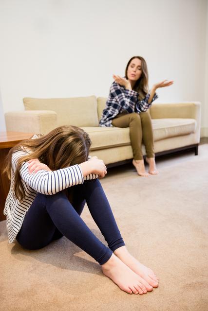 This image depicts a mother and daughter in a heated argument in a living room. The mother is sitting on a couch, gesturing with frustration, while the daughter is sitting on the floor with her head down, appearing upset. This image can be used to illustrate family conflicts, parenting challenges, emotional stress, and communication issues within a household. It is suitable for articles, blogs, and educational materials on family dynamics and mental health.