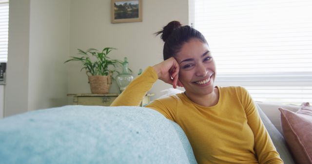 Smiling woman in a yellow sweater is relaxing on a light blue couch in a cozy living room. The room features houseplants, soft light, and comfortable furniture, creating a serene and relaxed atmosphere. This image is ideal for lifestyle blogs, home decor websites, mental health awareness campaigns, or advertisements focusing on comfort and relaxation.