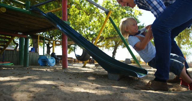 Mother helping blonde boy slide down playground slide while smiling on sunny summer day. Scene captures joy, parenting, and outdoor activities. Perfect for blogs, parenting magazines, promotional materials for parks, and family lifestyle articles.