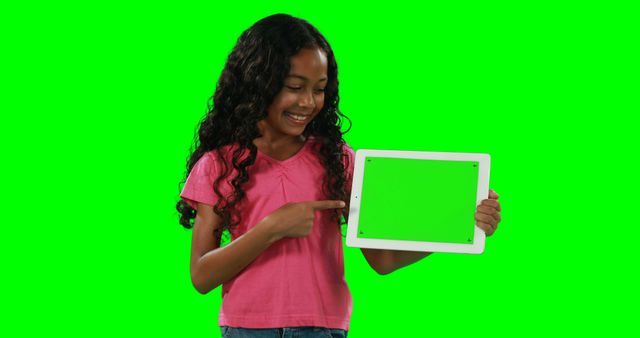 A young African American girl is smiling and pointing to a blank tablet screen, with copy space. Her cheerful expression and the green screen tablet invite viewers to imagine various educational or entertainment content.