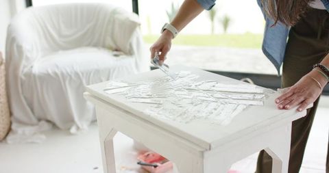 Mature woman refinishing wooden side table spreading white filler with putty knife