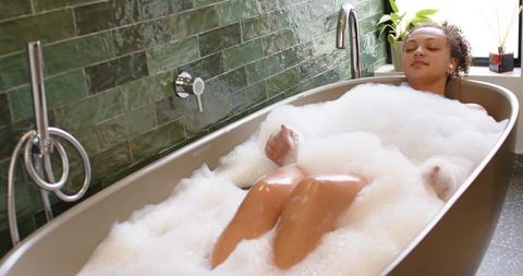 African american woman relaxing in bubble bath amid green-tiled bathroom with freestanding tub