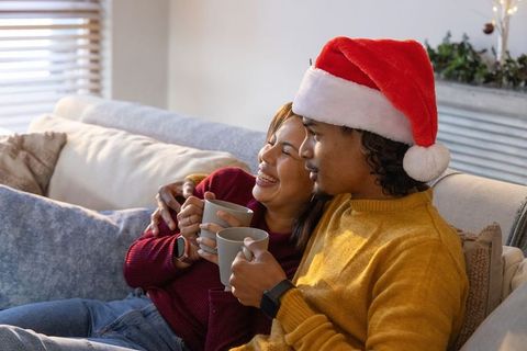 Diverse couple cuddling on sofa wearing santa hat and holding mugs, cozy christmas moment