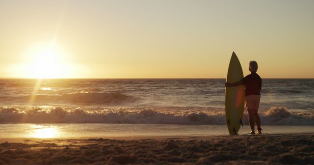 Surfer holding a surfboard watching sunset on beach. Suitable for themes involving surfing, leisure, summer, ocean activities, tranquility, and beach vacations.