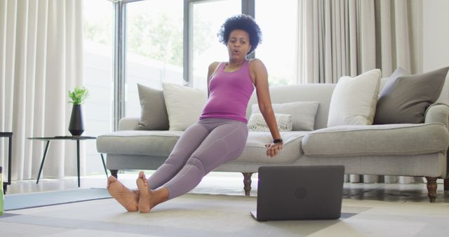 A woman is exercising at home, using an online fitness class. She is positioned in front of a laptop, engaging in a workout routine on a yoga mat in her living room. Natural light filters through large windows, creating a bright environment. Useful for promoting home workouts with online resources, fitness classes, healthy living, self-care, and indoor exercise routines.