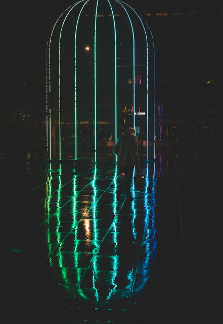 Person standing in front of glowing neon cage installation at night, reflecting off wet pavement. Ideal for illustrating modern urban nightlife, contemporary art installations, vibrant night scenes, and mood lighting.