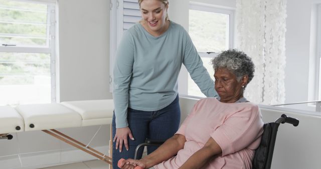 Senior woman sitting in wheelchair being assisted by a caregiver. The caregiver is providing support and encouraging physical therapy exercises. Ideal for use in healthcare promotions, lifestyle articles about elderly care, home therapy guides, and rehabilitation services.