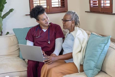 Home nurse assisting african american senior woman using laptop for telehealth and care
