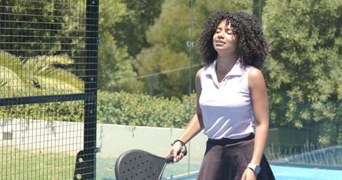 Woman playing padel on sunny court by pool holding racket wearing polo top and smartwatch