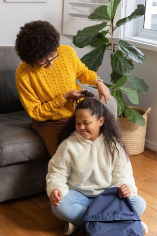 Caregiver styling child's hair while child smiles with backpack by sofa in living room