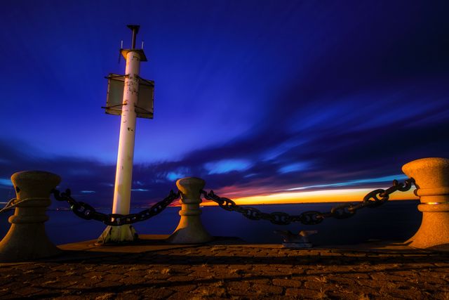 This scenic view captures a sunrise over the ocean, with deep blue skies and an orange glow on the horizon. The silhouettes of bollards and tugboat chains add an industrial nautical feel. Perfect for use in travel blogs, nautical-themed designs, inspirational artwork about new beginnings, or backgrounds depicting tranquility and peace.