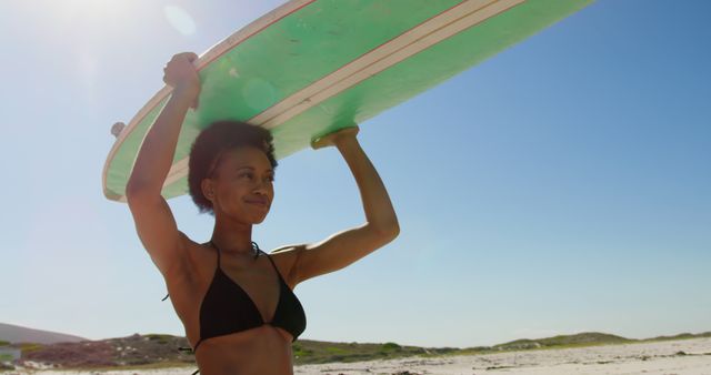 Young African American woman holds a surfboard on the beach, with copy space. She's ready for a day of surfing under the bright sun.