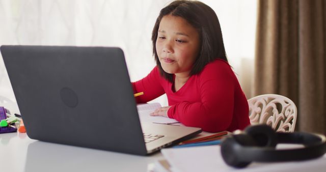 A young girl is engaged in online learning activities, working on schoolwork using a laptop at home. She appears focused while writing notes. This can be used for themes related to remote education, homeschooling, children using technology, and the impact of digital learning in modern education.