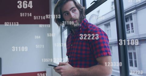 Bearded professional using smartphone by office window with floating numeric data overlay