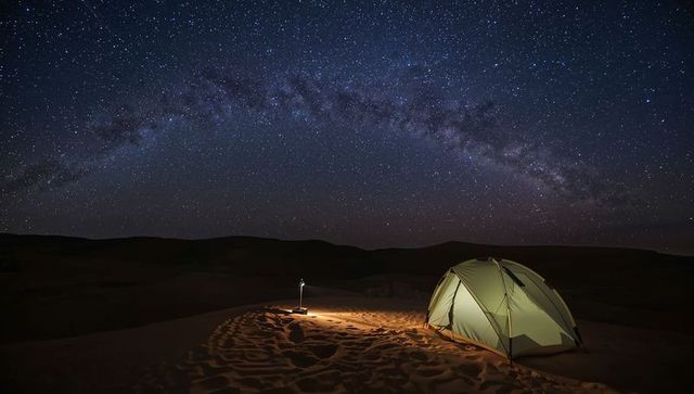 Camping under milky way in desert night with glowing tent and lantern, rippling sand