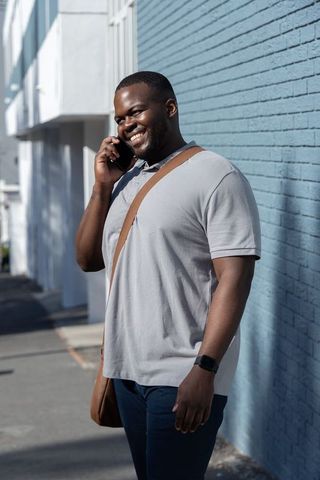 Smiling african american man talking on smartphone while standing by blue brick wall