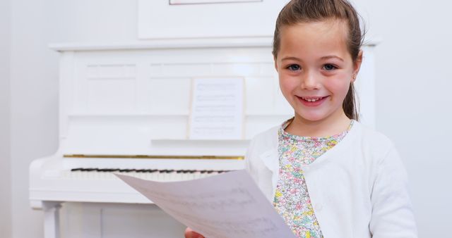 Little girl smiling while holding sheet music in a home setting with a white piano. Ideal for educational material, music learning content, or family-oriented advertisements.