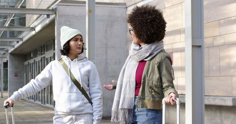 Two young travelers talking at urban building entrance with luggage under glass canopy