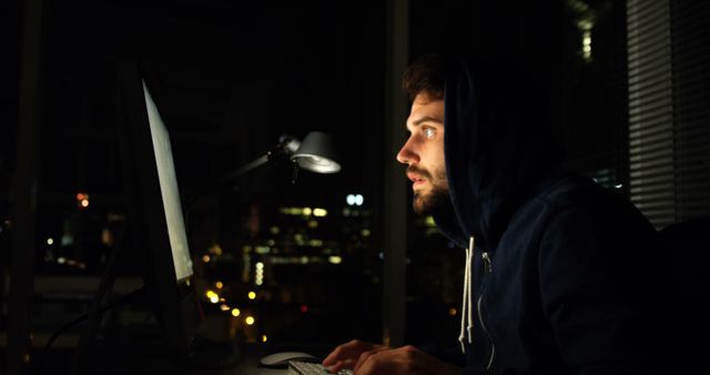 Hooded man working intensely on computer late at night. Useful for illustrating topics related to cybersecurity, hacking, programming, technology, and digital forensics. Great for articles, blog posts, and educational materials discussing internet security threats and hacking vigilance.