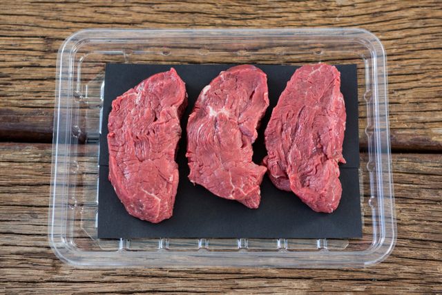 Three raw beef steaks in clear plastic packaging on rustic wooden table. Ideal for use in food blogs, grocery store advertisements, meal preparation guides, and butcher shop promotions.
