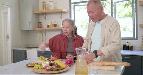 Senior couple preparing fruit platter and slicing baguette on sunlit kitchen island