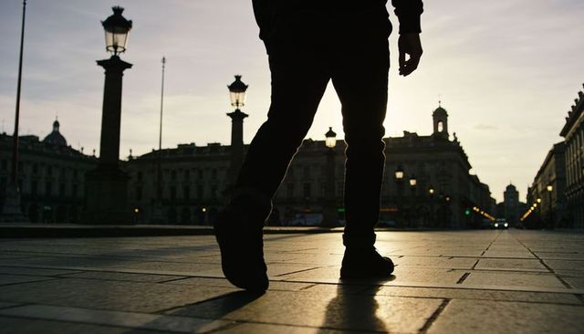 Backlit walking silhouette crossing historic city plaza at dusk with long shadow and lamps